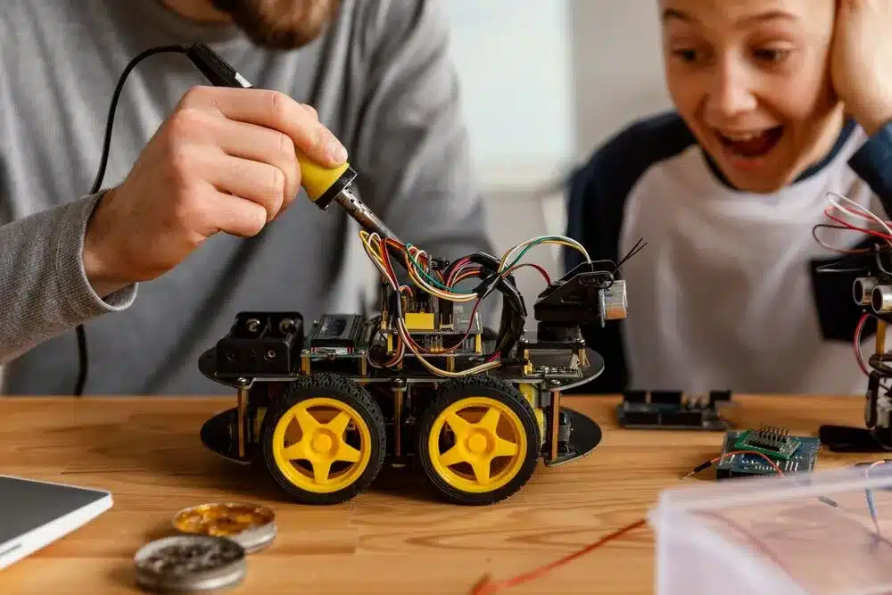 A teacher and student are testing a robot. The student is smiling and excited, and the teacher is watching with pride.